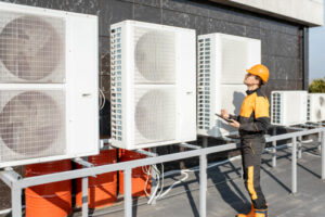 commercial hvac tech wearing black and yellow gear analyzing outdoor heating and cooling system on the roof of a commercial building during a sunny day