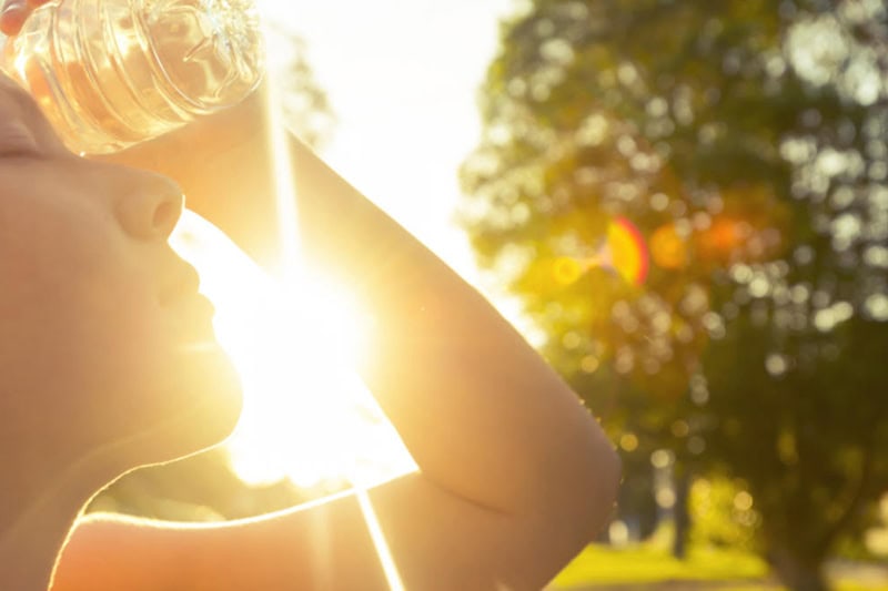 Prepare for Hot Weather With an Air Conditioning Tune-up. Photo of a woman trying to cool off in the hot sun using a cold water bottle.