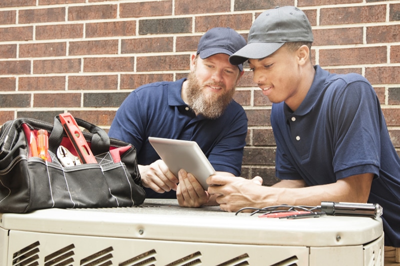 Multi-ethnic team of men repairing a home's air conditioner unit outdoors. They use a digital tablet to access the repairs. They both wear blue uniforms.
